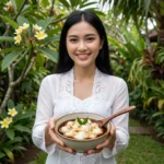 A smiling Indonesian woman wearing a modern kebaya holding a bowl of creamy bubur sumsum in a lush tropical garden setting.