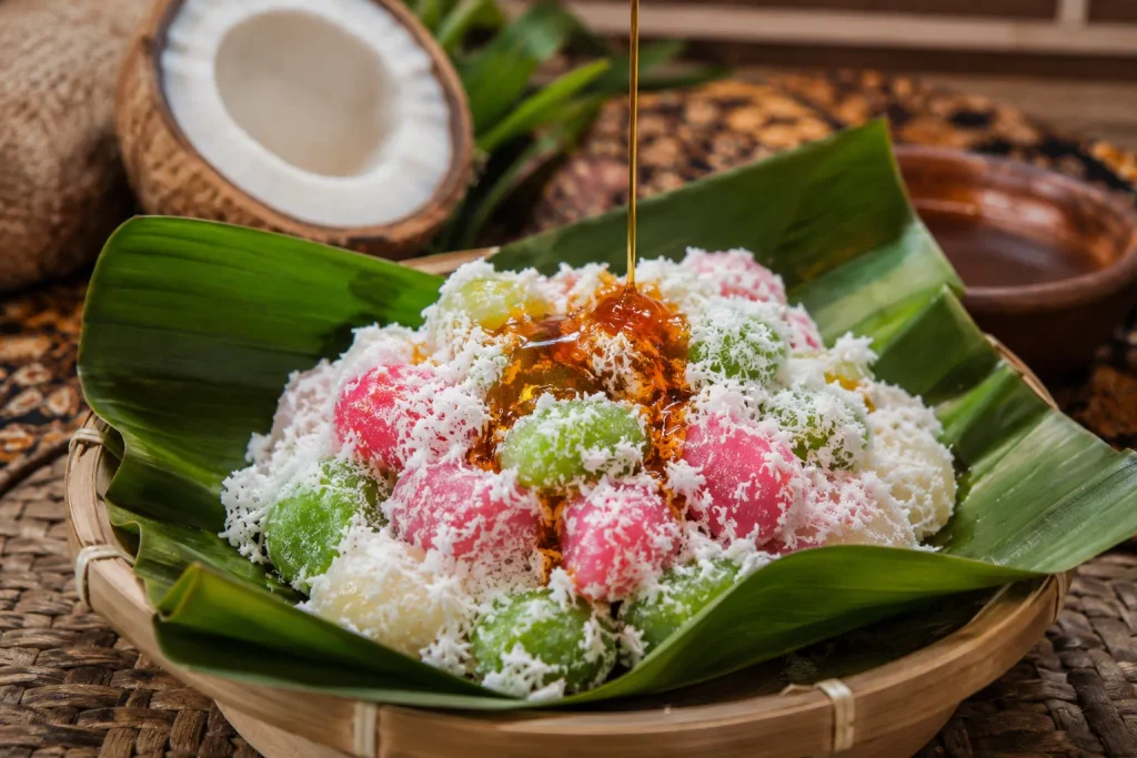 Traditional Indonesian cenil dessert served on a fresh banana leaf, topped with grated coconut and glossy palm sugar syrup in a warm kitchen setting.